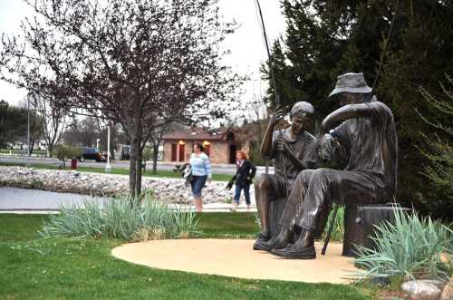 A bronze statue of two fishermen sitting on a bench, surrounded by greenery and a walking path near a pond.