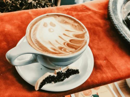 A cup of coffee with ghost latte art sits on a saucer, accompanied by a small piece of cake.