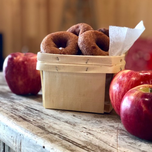 A wooden basket filled with donuts, surrounded by fresh red apples on a rustic wooden table.
