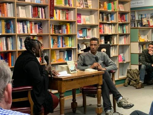 A woman and a man sit at a table in a bookstore, engaged in conversation, with bookshelves in the background.