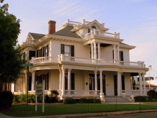 A large, historic white Victorian house with multiple balconies and green shutters, set against a clear sky.