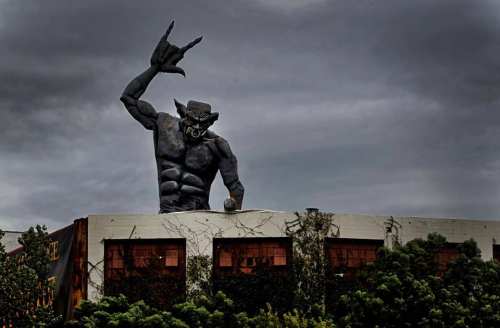 A large, menacing statue of a muscular figure with horns, raising a hand in a rock gesture against a cloudy sky.