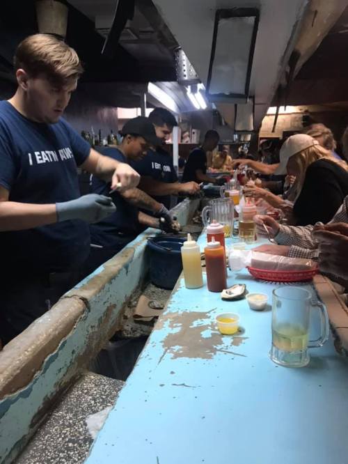 A busy bar scene with patrons enjoying drinks and food, while staff prepare seafood at a counter.