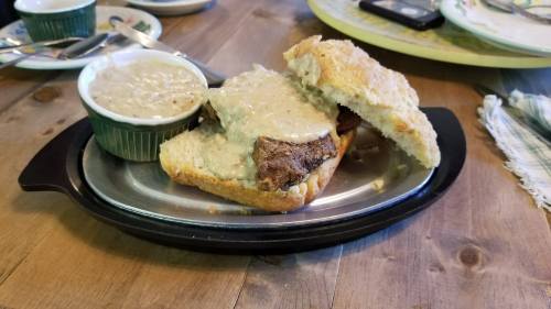 A hearty sandwich with meat and creamy sauce, served on a metal plate alongside a bowl of soup.