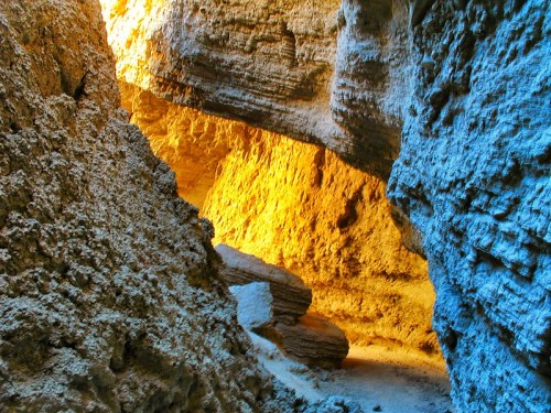 A narrow canyon with textured rock walls illuminated by warm orange and cool blue light.