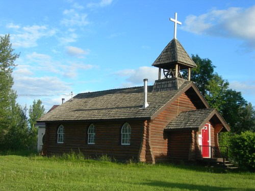 A small wooden church with a red door and a bell tower, surrounded by greenery under a blue sky.
