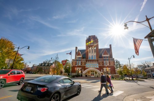 A sunny day in a town square with a historic building, two pedestrians crossing, and cars parked nearby.