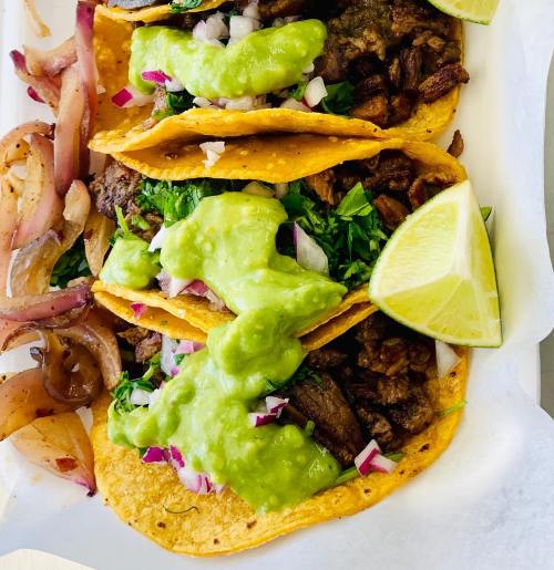 Three tacos filled with beef, topped with guacamole, onions, and cilantro, served with grilled onions and a lime wedge.
