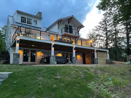 A modern two-story house with a large deck, surrounded by trees, under a cloudy sky at dusk.