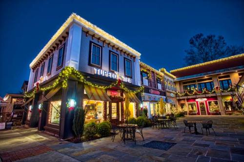 A charming storefront decorated with holiday lights, surrounded by greenery, at dusk. Cozy outdoor seating is visible.