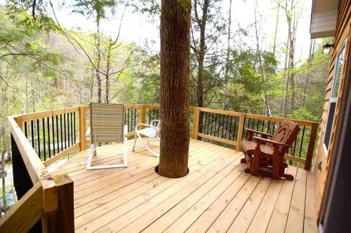 A wooden deck surrounded by trees, featuring a chair and a lounge chair, with a tree growing through the center.