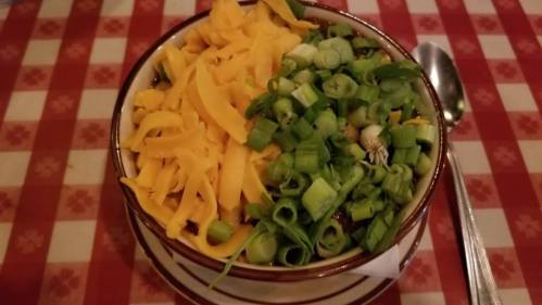 A bowl filled with shredded cheddar cheese and chopped green onions on a checkered tablecloth.