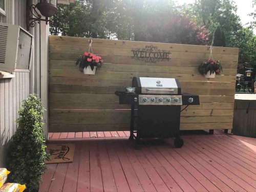 A black grill on a wooden deck with a welcome sign and hanging flower pots in a backyard setting.