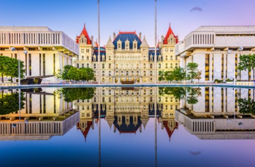 New York State Capitol building reflected in a calm pool, surrounded by modern architecture and trees at sunset.