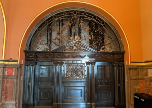 Ornate black door with intricate metalwork, set against a warm orange wall in a historic building.