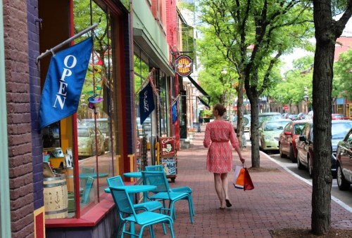 A woman walks down a vibrant street lined with shops, trees, and colorful chairs, carrying a shopping bag.