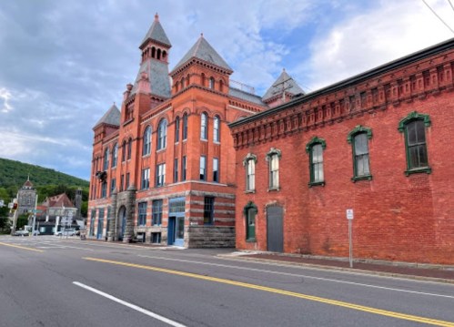 Historic red brick building with towers, set against a cloudy sky, alongside a quiet street.