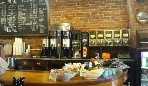 A cozy café counter with a menu board, coffee machines, and a display of baked goods and snacks.