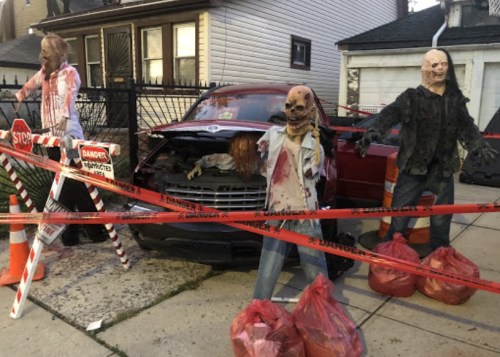 A spooky Halloween display with zombie figures near a car, surrounded by caution tape and bags.