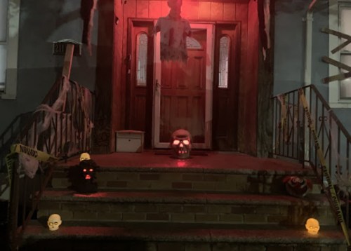 A spooky Halloween scene featuring a decorated front porch with a glowing skull, ghostly figure, and eerie lighting.