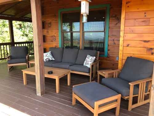 A cozy porch with wooden furniture, including a sofa, chairs, and a coffee table, surrounded by nature.