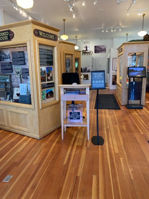 Interior of a museum with wooden floors, display cases, and informational signs. A welcome desk is visible in the foreground.