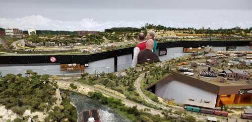 A detailed model train layout featuring landscapes, buildings, and a river, with two people observing the scene.