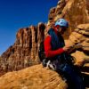 A climber in a helmet sits on a rock, checking their gear against a backdrop of rugged cliffs and blue sky.