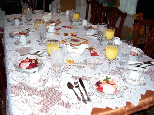 A beautifully set dining table with lace tablecloth, plates of fruit, glasses, and decorative autumn leaves.