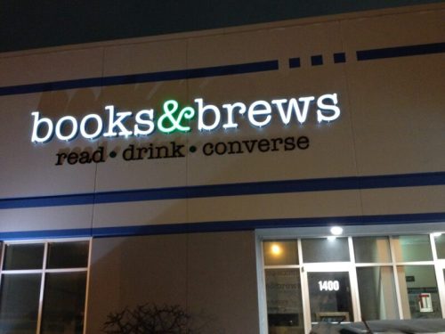 Sign for "books & brews" featuring the tagline "read drink converse" on a building exterior at night.