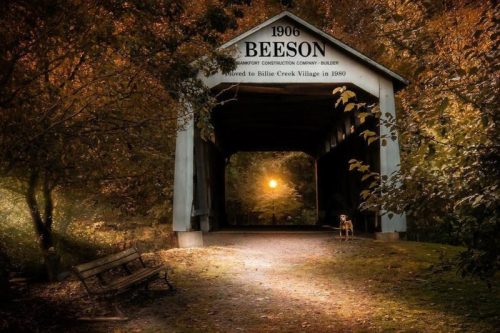 A covered bridge surrounded by autumn foliage, with a bench and a dog in the foreground, illuminated by a sunset.