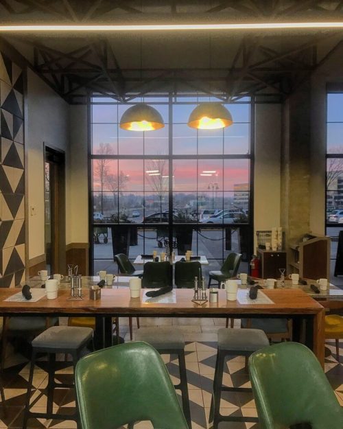 A cozy restaurant interior with a wooden table, green chairs, and a view of a colorful sunset through large windows.