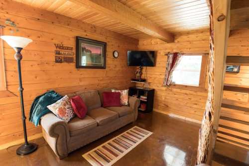Cozy wooden cabin interior featuring a sofa, decorative pillows, a lamp, and a TV. Warm, inviting atmosphere.
