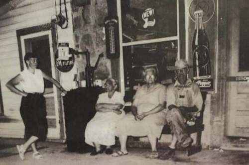 A black-and-white photo of four people sitting on a bench outside a store, with vintage signs in the background.