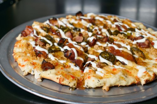 A close-up of a pizza topped with bacon, jalapeños, and drizzled with white sauce on a metal tray.