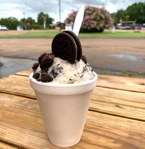 A cup of ice cream topped with crushed Oreos and a whole Oreo cookie, sitting on a wooden table outdoors.