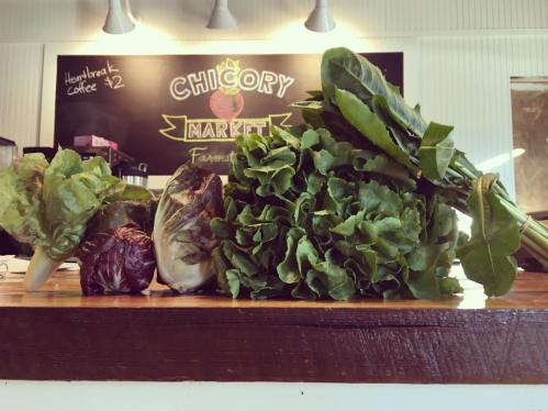 A variety of fresh greens and lettuce displayed on a wooden counter, with a chalkboard menu in the background.