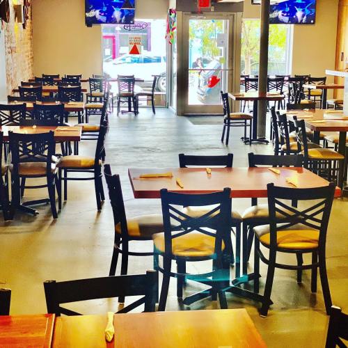 A spacious restaurant interior with empty tables and chairs, featuring brick walls and large windows.