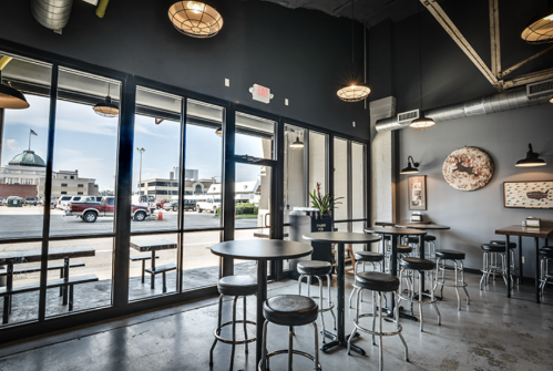 Modern café interior with wooden tables, metal stools, and large windows showcasing an outdoor view.