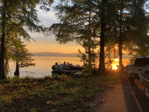 Sunset over a lake with silhouettes of people on a dock, surrounded by trees and a parked vehicle nearby.