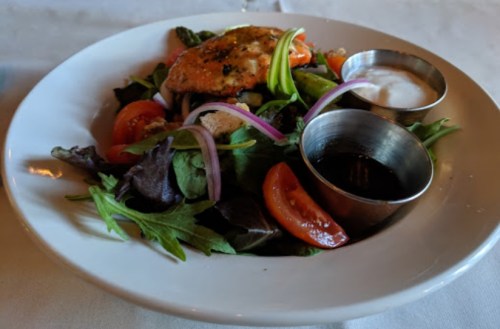 A fresh salad with mixed greens, tomatoes, red onions, and a piece of salmon, served with two dressings on the side.