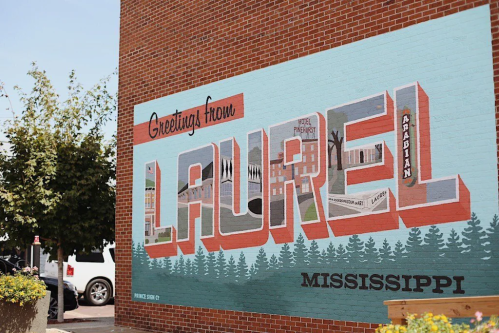 A colorful mural reading "Greetings from Laurel, Mississippi" with trees and buildings in the background.