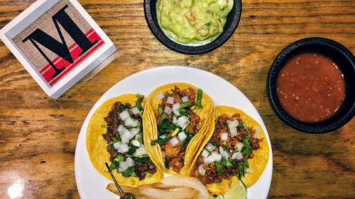 Three tacos with meat, onions, and cilantro on a plate, accompanied by guacamole and beans on a wooden table.