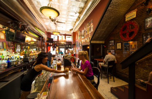 A lively bar scene with a bartender serving drinks, patrons at the bar, and vintage decor on the walls.