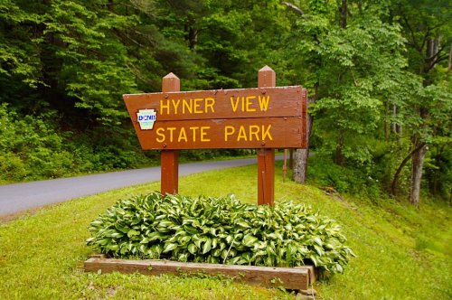 Wooden sign pointing to Hyner View State Park, surrounded by greenery and a road in the background.