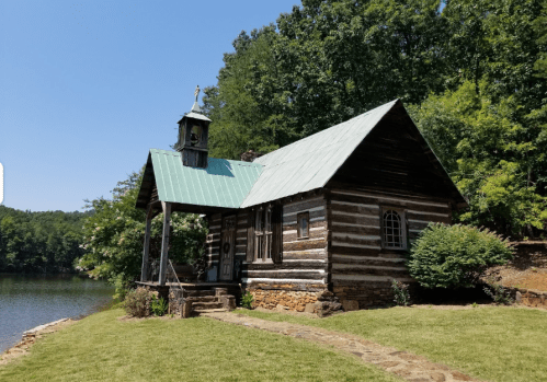 A rustic log cabin with a green metal roof, situated by a serene lake and surrounded by lush greenery.
