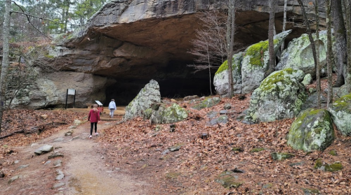 Two people walk towards a large rock cave surrounded by trees and scattered rocks, with autumn leaves on the ground.