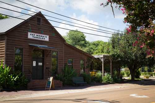 A rustic wooden building with a sign reading "Fat Mama's Tamales," surrounded by greenery and a clear blue sky.