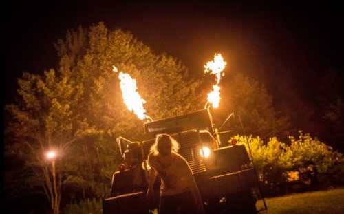 A person stands in front of a truck with flames shooting from its roof, set against a dark, wooded background.