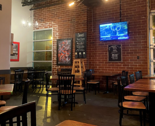 A dimly lit restaurant interior with wooden tables, chairs, and a TV displaying news on a brick wall.